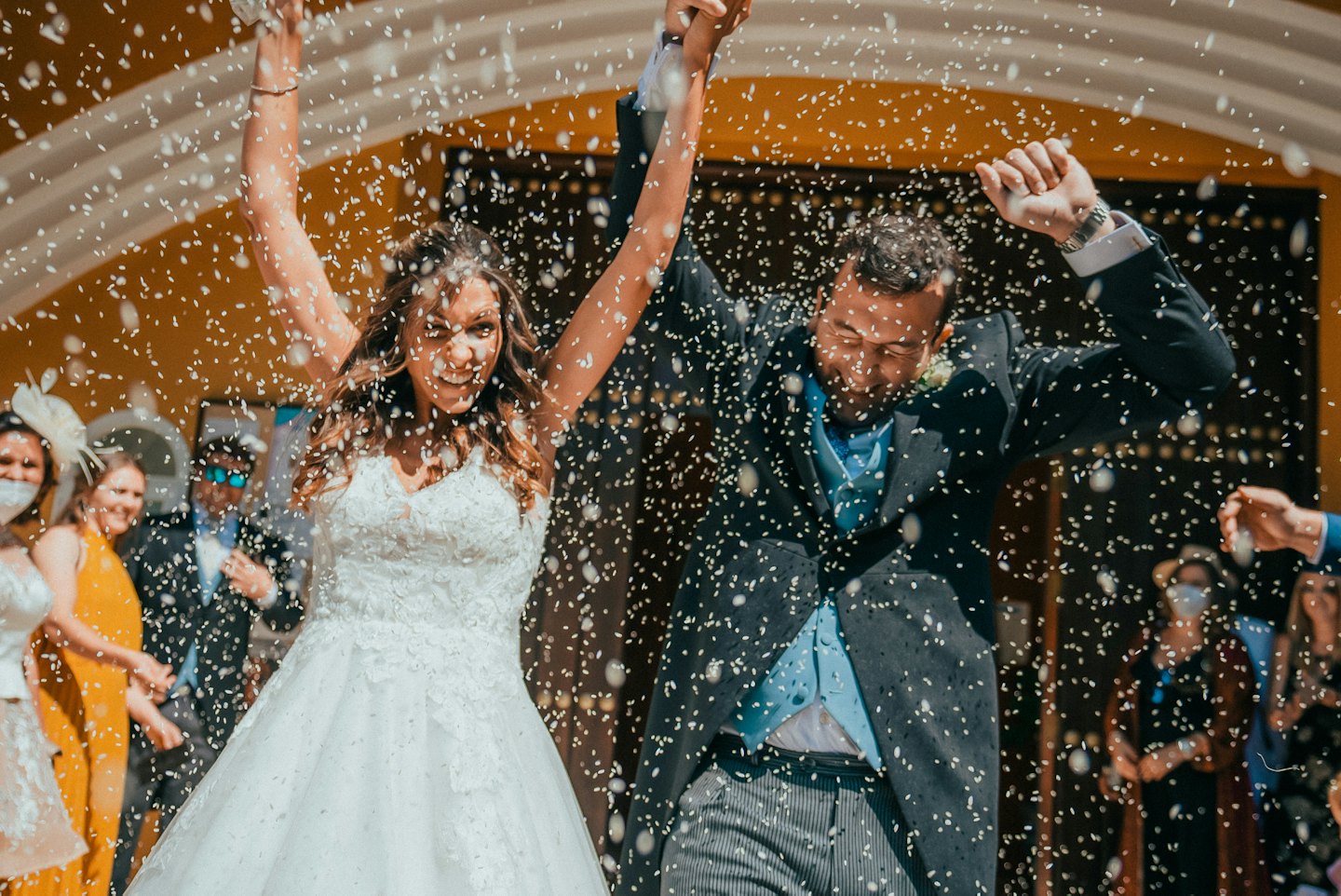 Bride and groom celebrate with arms raised in confetti.